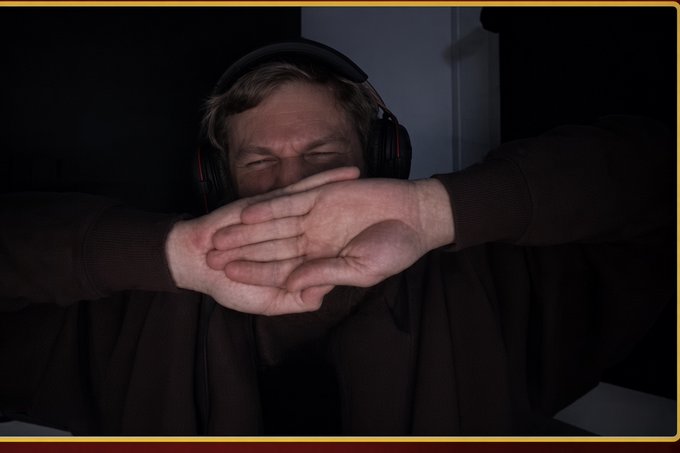 A man wearing headphones covers his face with both hands in a dark room, possibly reacting to something shocking or emotional.