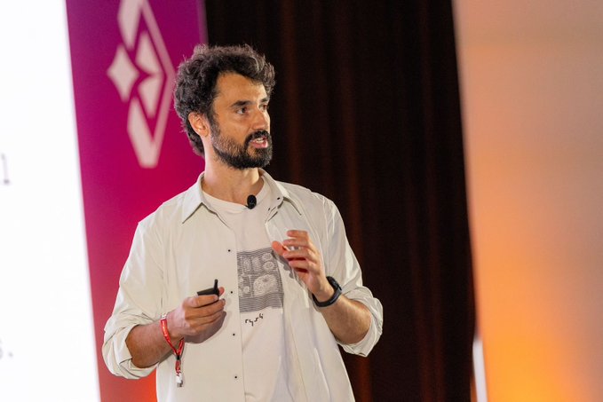 The image shows a man standing on stage, giving a speech or presentation. The background features a large screen displaying text related to the future of volatility markets. The speaker appears to be discussing the potential impact of institutions bringing assets to blockchain networks and the need for options and derivatives infrastructure in these new market environments.