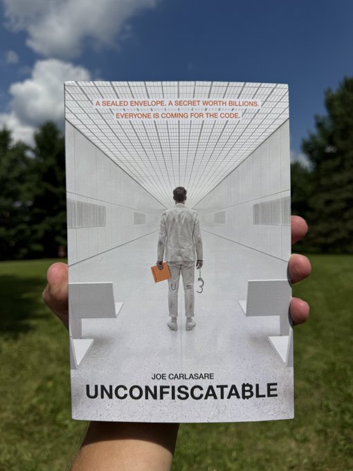 A hand holds up a paperback book titled 'UNCONFISCATABLE' by Joe Carlasare outdoors; the cover shows a person in white walking down a corridor with text reading 'A SEALED ENVELOPE. A SECRET WORTH BILLIONS. EVERYONE IS COMING FOR THE CODE.'