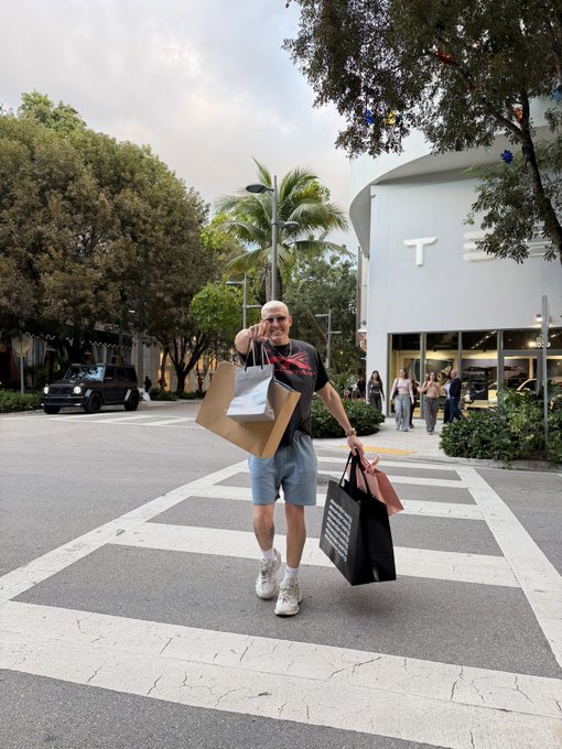 A man wearing a black graphic tee, denim shorts, and sunglasses stands in a crosswalk holding multiple shopping bags near a Tesla store, giving a peace sign.