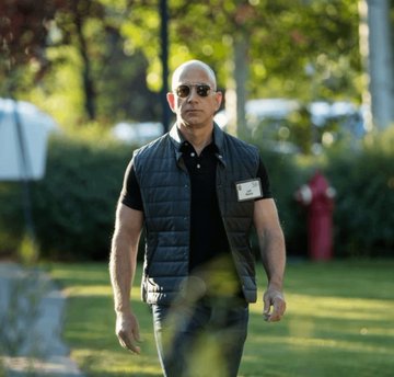 Jeff Bezos, wearing sunglasses and a navy quilted vest with a visible name badge, walks outdoors, accompanying news of his filing to sell up to 15 million Amazon shares worth approximately $4.07B.