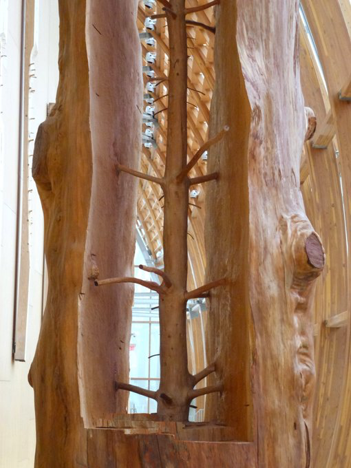 A close-up photograph of a wooden sculpture featuring a carved tree-like branch structure enclosed between two rough-hewn wooden slabs in what appears to be a timber-framed building.