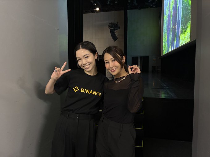 Two smiling women pose at the Japan Blockchain Week Summit, with one wearing a black "BINANCE" t-shirt. They appear to be event staff or attendees welcoming the opening of #JBWS2026, standing in a venue with a large screen displaying forest imagery in the background.