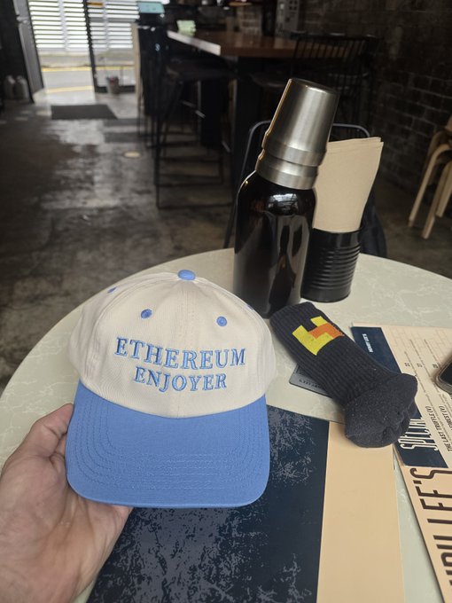 A hand holds up a cream and blue baseball cap with 'ETHEREUM ENJOYER' embroidered in blue text, displayed on a marble table alongside a dark cocktail shaker and rolled napkins.