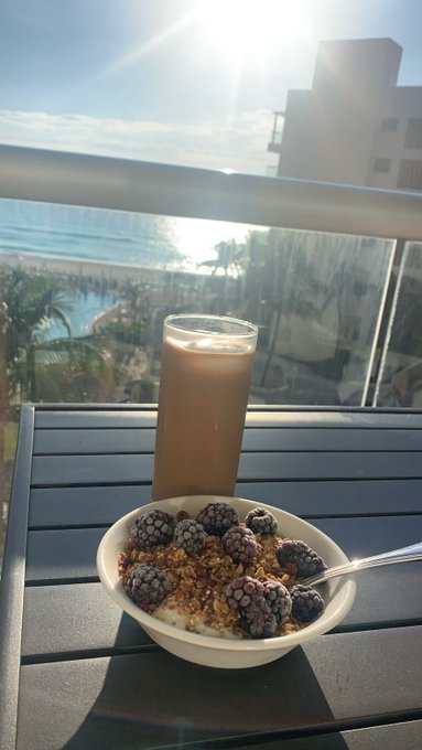 A tropical breakfast spread featuring a bowl of yogurt, blackberries, and granola alongside a glass of iced coffee sits on a balcony overlooking a sunlit ocean view—reflecting the relaxed, optimistic "GM" (good morning) vibe of the post celebrating $ANSEM holders and Bullpen traders amid the market's upward momentum.