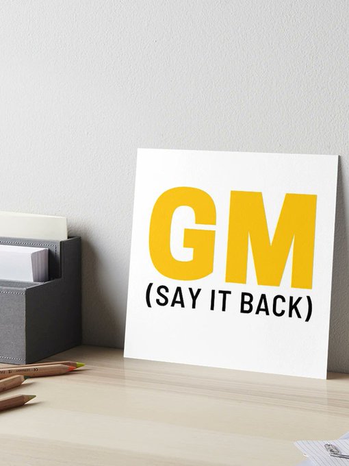 The image features a white sign that reads "G.M." written in yellow letters, placed on top of a wooden desk or table. The sign also has a message about saying "it back" written below it. There are two pencils lying next to the sign and a book is positioned nearby as well.