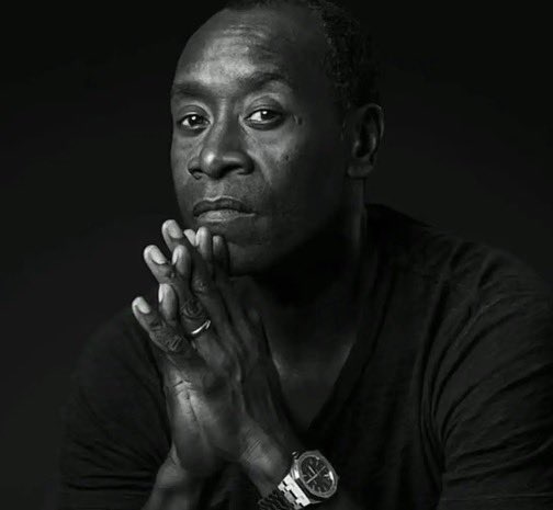 A black-and-white portrait of a man with his hands clasped together near his chin, wearing a ring and a wristwatch.