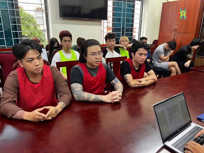 A group of tattooed young men wearing red and yellow vests sit at a conference table in front of a laptop in what appears to be an office or institutional setting.