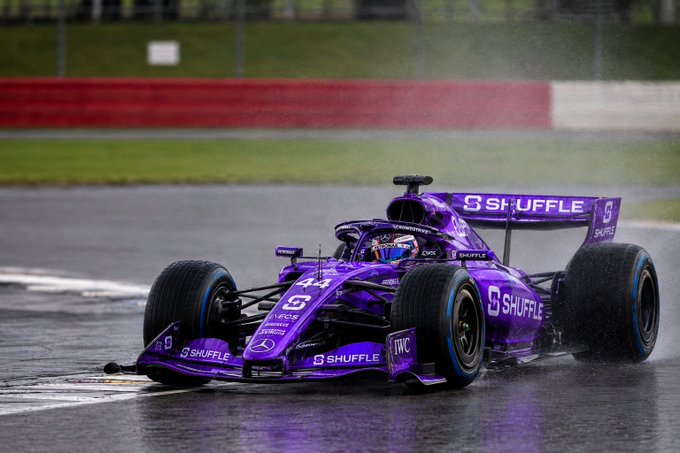 A purple Formula 1 race car with 'SHUFFLE' branding and the number 44 speeds along a wet track, kicking up spray from its tires.