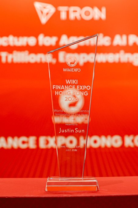 A crystal award from Wiki Finance Expo Hong Kong 2025 sits on display, engraved with "World's Most Influential Innovator of the Year" presented to Justin Sun. The backdrop features the TRON logo and text referencing an AI infrastructure, directly tying to the post's acknowledgment of WikiExpo's recognition of TRON's innovative work alongside @BAI_AGI.