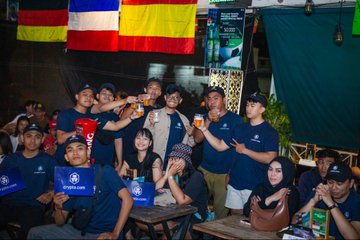 A group of people posing together at what appears to be a fan event, with flags (German, French, Spanish) visible in the background and many wearing matching blue shirts.