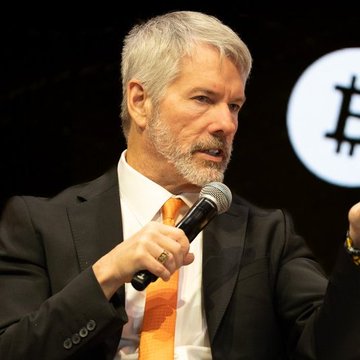 A man with gray hair and a beard, wearing a dark suit and orange tie, speaks into a microphone at what appears to be a Bitcoin event, with a Bitcoin logo visible in the background—visually reinforcing Michael Saylor's bold statement that "The world will be built on Bitcoin."