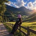 A scenic landscape photo of a person wearing a pink beanie leaning against a wooden fence, with green hills and a bright sky in the background.