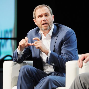 A man in a blue suit and white shirt, likely Brad Garlinghouse, gestures expressively while speaking during what appears to be a panel discussion or conference. His animated hand gestures suggest he is making a pointed argument, aligning with his critical comments on Michael Saylor's Bitcoin funding model and its impact on the crypto market.