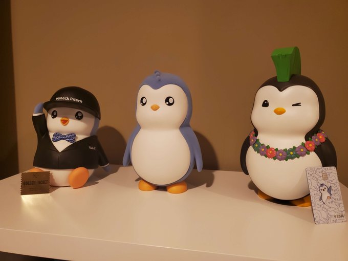 Three collectible penguin figurines sit on a shelf, including one wearing a 'peacock intern' cap with a blue bow tie, a plain blue and white penguin, and one wearing a green leafy headpiece with a flower lei, accompanied by a 'Golden Ticket'.