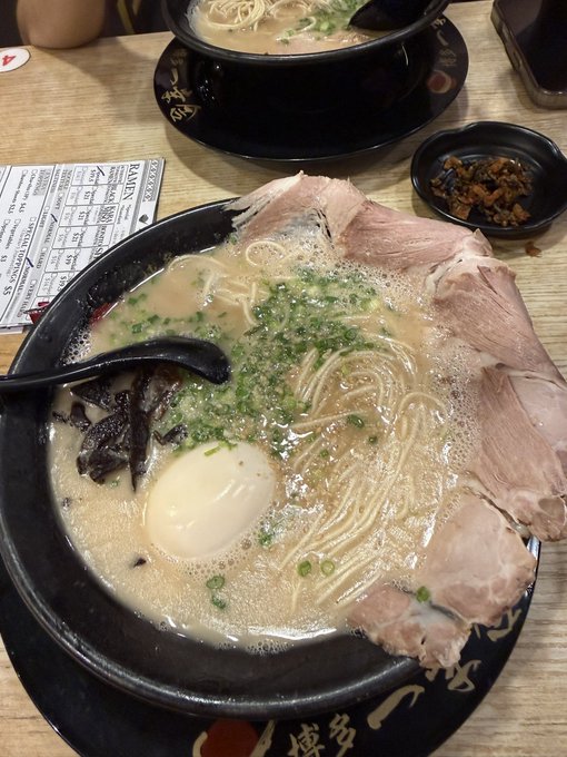 A bowl of tonkotsu ramen with chashu pork, a soft-boiled egg, nori, and green onions, served on a black tray with a side dish of pickled vegetables.