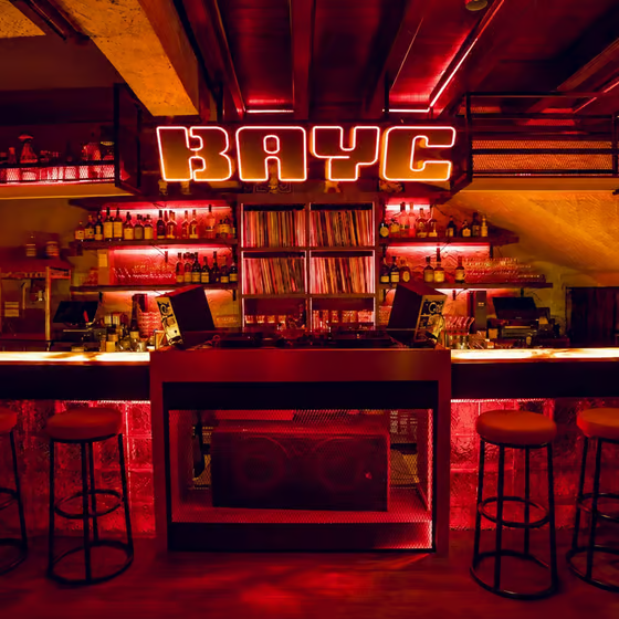 A bar interior with red neon lighting and a prominent "BAYC" (Bored Ape Yacht Club) illuminated sign, with vinyl records on shelves and bottles behind the counter.