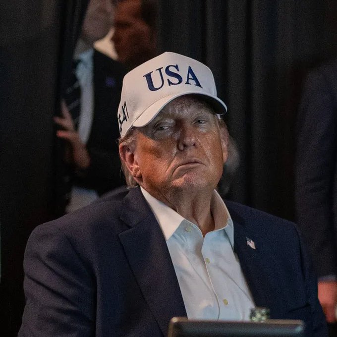 A man wearing a white "USA" cap and a navy blazer over a white shirt sits with a serious expression in what appears to be an indoor event setting.