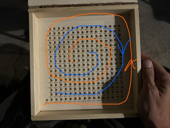 A hand holds a wooden box filled with drilled holes containing handwritten numbers, overlaid with orange and blue spiral drawings illustrating the double spiral numbering system described. The caption highlights how this offset spiral pattern neutralizes mechanical bias, working in tandem with a base 257/mod 2048 extraction method to generate "whole entire entropy."