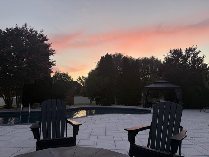 Two Adirondack chairs face a serene pool and gazebo at sunset, with soft pink and orange hues painting the sky above the tree line—a peaceful scene inviting viewers to step outside and enjoy nature's beauty.