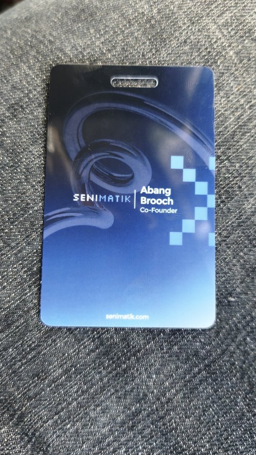A blue lanyard badge lies on a textured fabric surface, reading "SENIMATIK | Abang Brooch | Co-Founder."