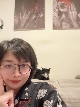 A person with glasses takes a selfie in front of a wall with artwork, while a black and white cat rests on a bed behind them.