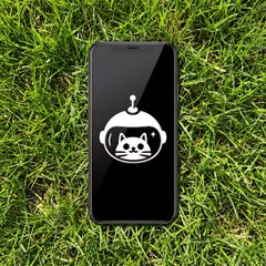 A black smartphone lies on green grass displaying a cartoon white cat logo wearing an astronaut helmet on its screen.