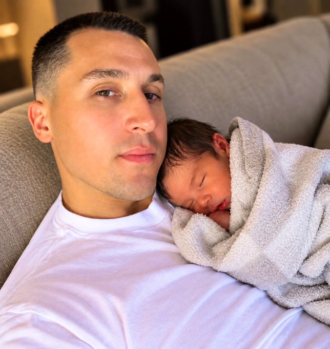 Intimate portrait of a young man in a white shirt resting on a couch while holding a sleeping newborn baby wrapped in a gray towel.