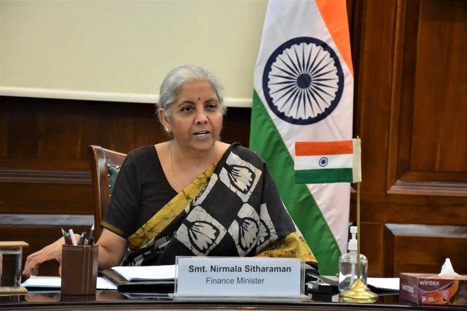 Indian Finance Minister Smt. Nirmala Sitharaman seated at a desk in front of the Indian flag, with a nameplate reading 'Smt. Nirmala Sitharaman, Finance Minister'.