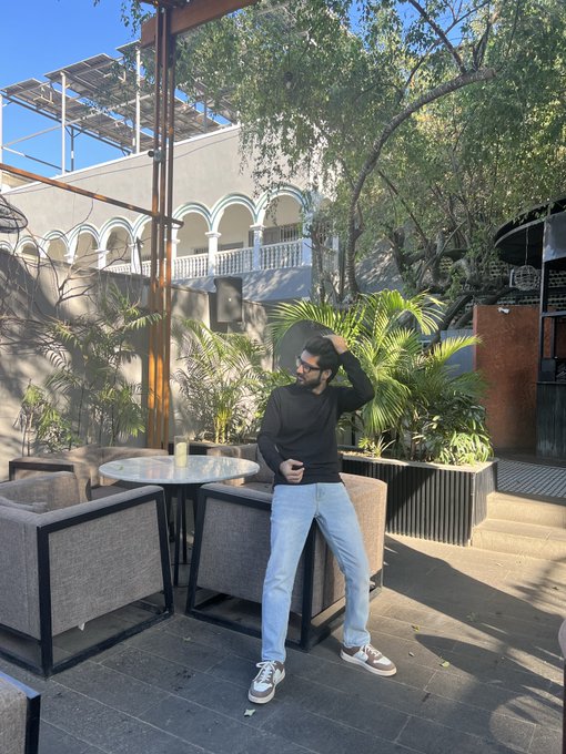A man in a black sweater and light blue jeans stands in an outdoor patio area with greenery, a table, chairs, and a building with arched architecture in the background.
