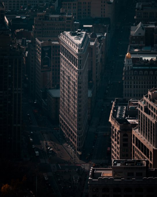 An aerial view of the iconic Flatiron Building in New York City, bathed in shadow among other surrounding city blocks.