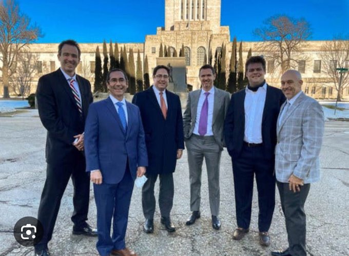 The image features a group of six men standing together, all dressed in suits and ties, posing for a photo. They are positioned in front of a large building with columns, which appears to be a courthouse or some other significant public structure. The men seem to be celebrating an accomplishment related to the landmark they are standing in front of.