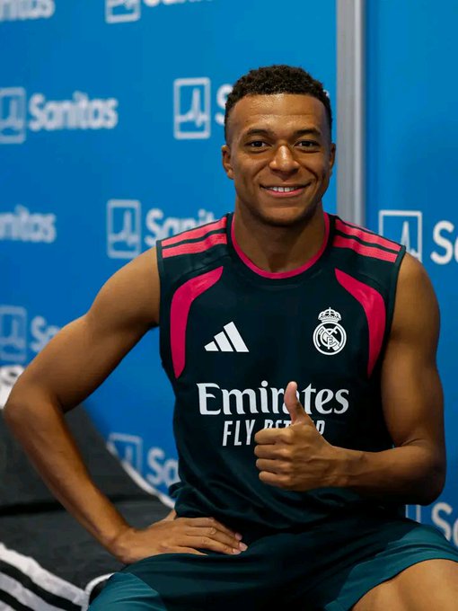 A smiling young man in a black and pink Real Madrid sleeveless training top with 'Emirates FLY BETTER' text gives a thumbs-up in front of a blue 'Sanitas' backdrop.