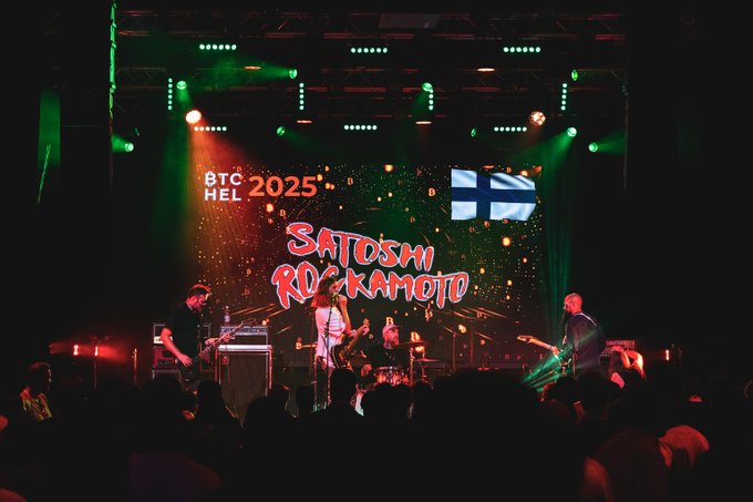 A concert photograph showing a live performance at 'BTC 2025 HEL' featuring 'Satoshi Rockamoto' displayed on a large screen, with green stage lighting and musicians performing for a crowd.
