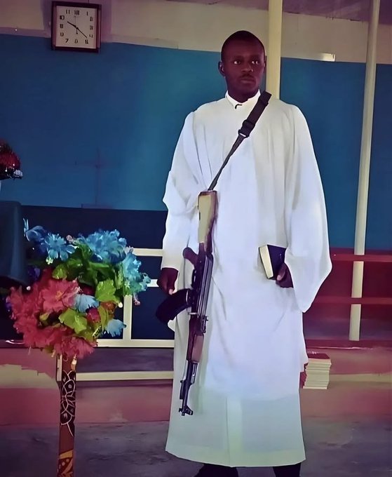 A man in a white religious robe stands holding a rifle and a small black book, with colorful flowers in the foreground.