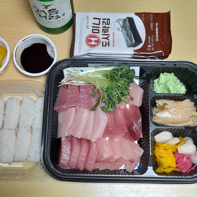 A takeout meal featuring sliced raw fish (sashimi) sushi rolls, wasabi, ginger, and pickled vegetables, accompanied by Korean seaweed snacks and a bottle of soju.