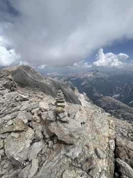 A photograph from a mountain summit showing a small stack of rocks (cairn) in the foreground with clouds and distant mountain ranges in the background.