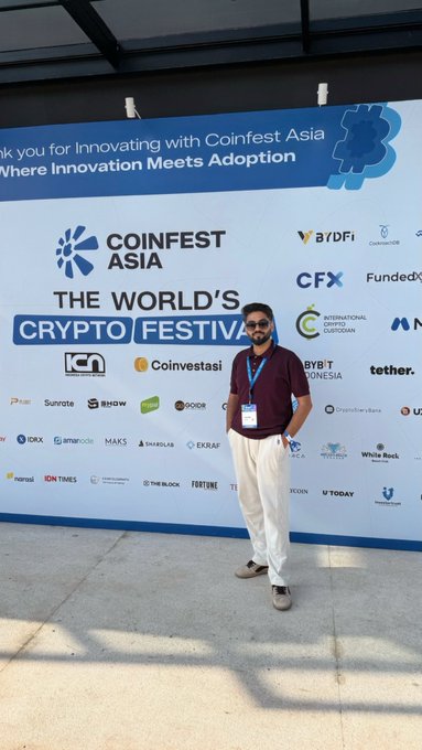 Photo of a man wearing sunglasses, a maroon polo shirt, white pants, and a lanyard posing in front of a Coinfest Asia sponsor backdrop reading 'THE WORLD'S CRYPTO FESTIVAL' with logos including ICH, ABDFi, CFX, FundEx, Coinvestasi, tether, and others.