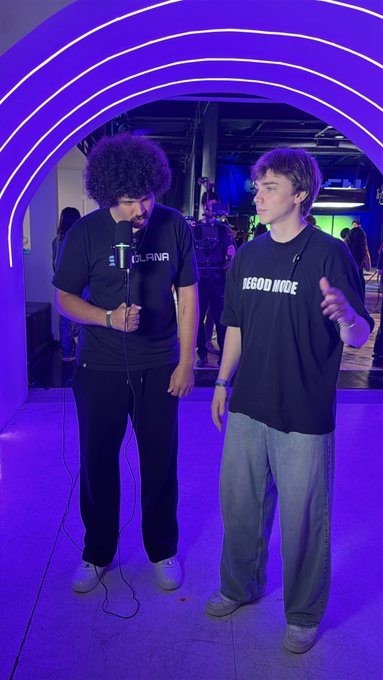 Two men standing in an archway illuminated with purple neon lights. The man on the left wears a black 'LOLANA' t-shirt and holds a microphone, while the man on the right wears a black 'WGOOD MODE' t-shirt.