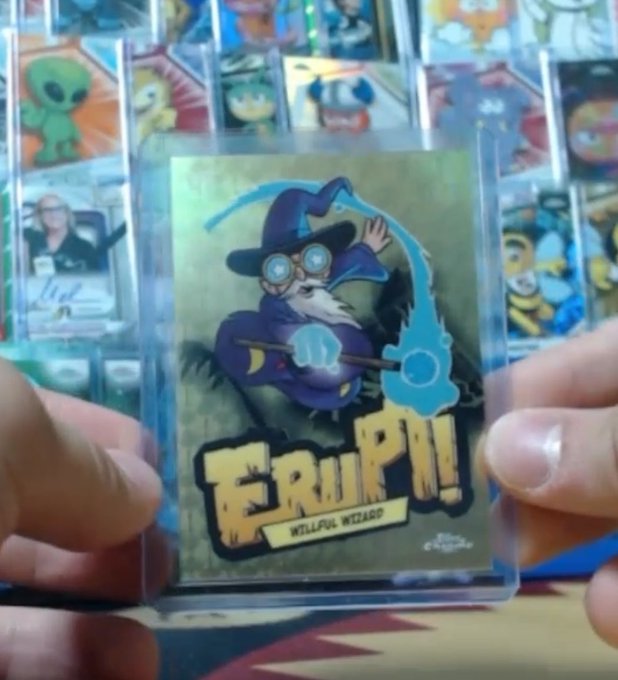 Hands hold up a trading card featuring a wizard character named 'FRUPI! - WILLFUL WIZARD' in a clear protective case, with a backdrop of other cartoon collectible cards.