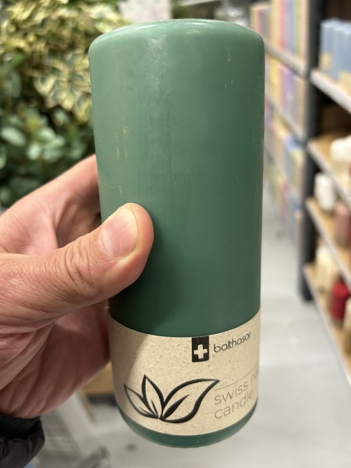 A hand holding a dark green cylindrical candle with a label reading 'bathospor' and 'swiss made candle' at what appears to be a retail store with shelves of products in the background.