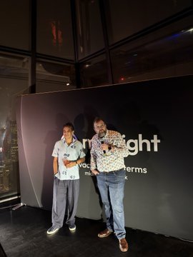 Two men pose together in front of a dark backdrop with partial text reading 'ght' and 'terms', with one wearing a white polo and the other in a patterned jacket.