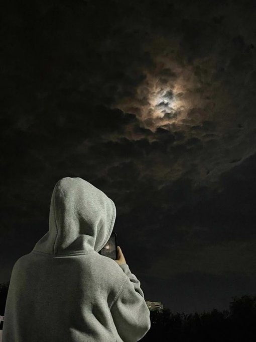 A person in a gray hoodie stands under a cloudy night sky, photographing the moon through their phone.