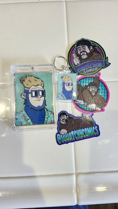 A collection of merchandise featuring "SQUATCH COMICS" patches and keychains, including a character with a beard and sunglasses in a tropical shirt.