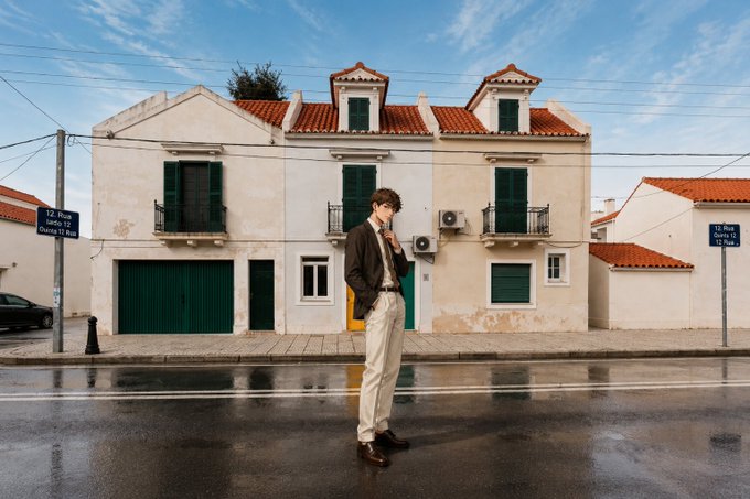 A young man in a brown jacket and light pants stands in the street in front of a Mediterranean-style building with green shutters and red roof tiles, with street signs reading "12. Rua Serpa Pinto" and "13. Rua Padre L.J. Roca".