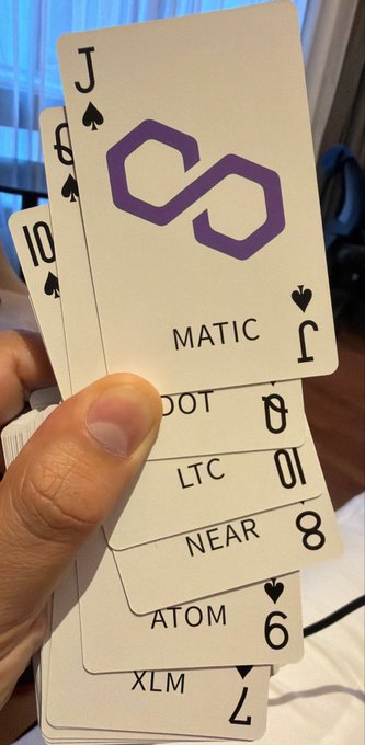 A hand holding a fan of playing cards themed after cryptocurrencies, showing MATIC (Jack), DOT, LTC, NEAR, ATOM, and XLM cards.