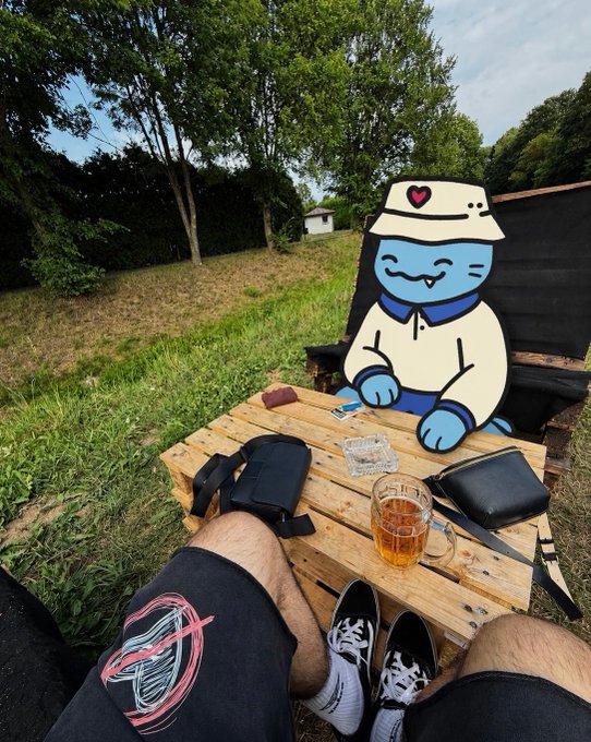 A first-person view of someone sitting outdoors at a wooden table with a glass of iced drink, shoes, and bags, with a cartoon blue cat character wearing a bucket hat superimposed in the chair across from them.