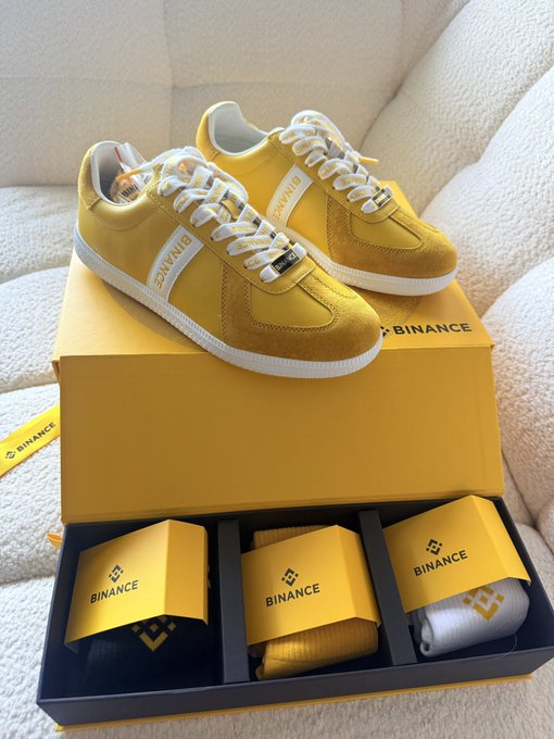 A pair of yellow and white Binance-branded sneakers displayed on top of yellow Binance shoe boxes, with additional socks and accessories in matching Binance packaging.