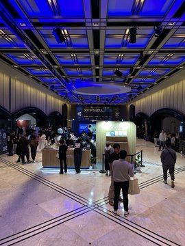 A large, modern event hall with a striking blue-lit ceiling is filled with attendees walking around exhibition booths, including one labeled "XP FINTECH."