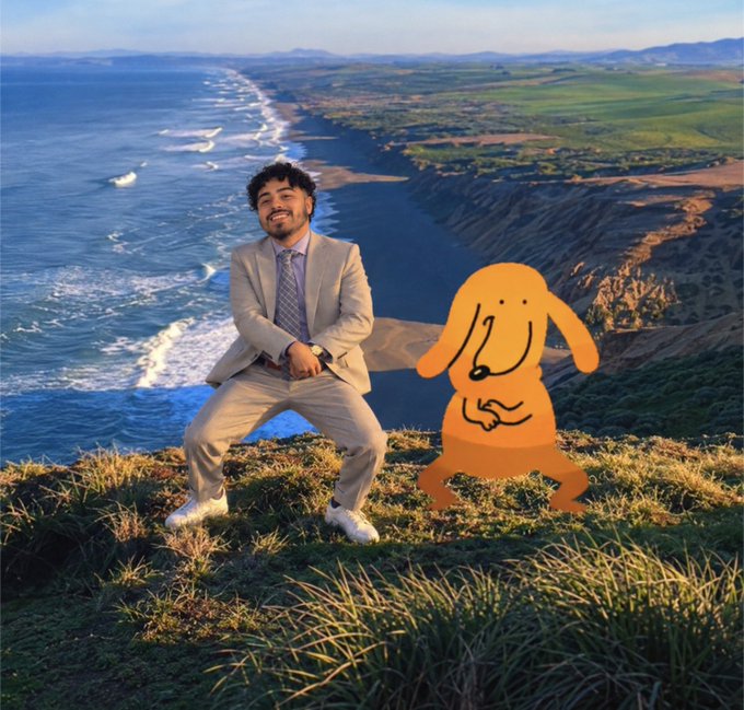 A man in a beige suit posing in a wide-legged stance alongside an orange cartoon rabbit character on a grassy hilltop overlooking a scenic coastline with cliffs and ocean.